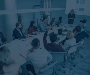 A group of people sit around a conference table watching a presenter give an ERP-focused presentation with charts projected on a screen.