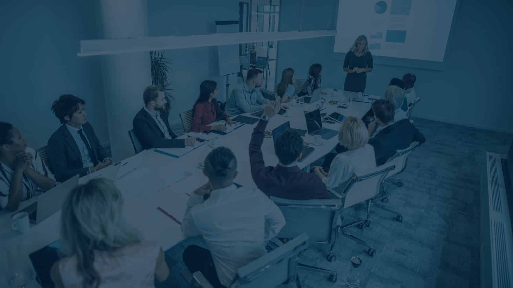A group of people sit around a conference table watching a presenter give an ERP-focused presentation with charts projected on a screen.