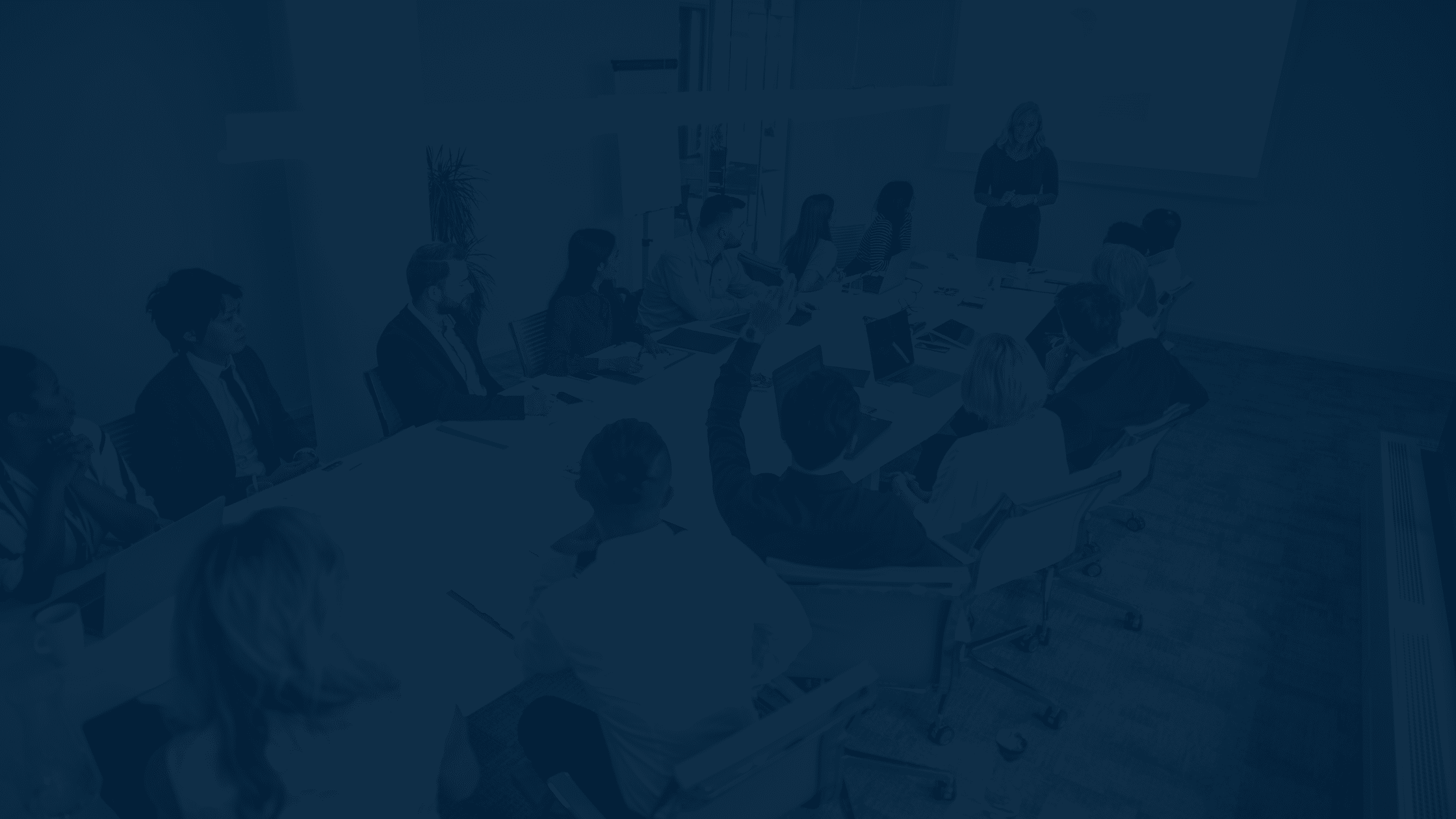 A group of people sit around a conference table while a person stands in front of a screen giving a presentation.