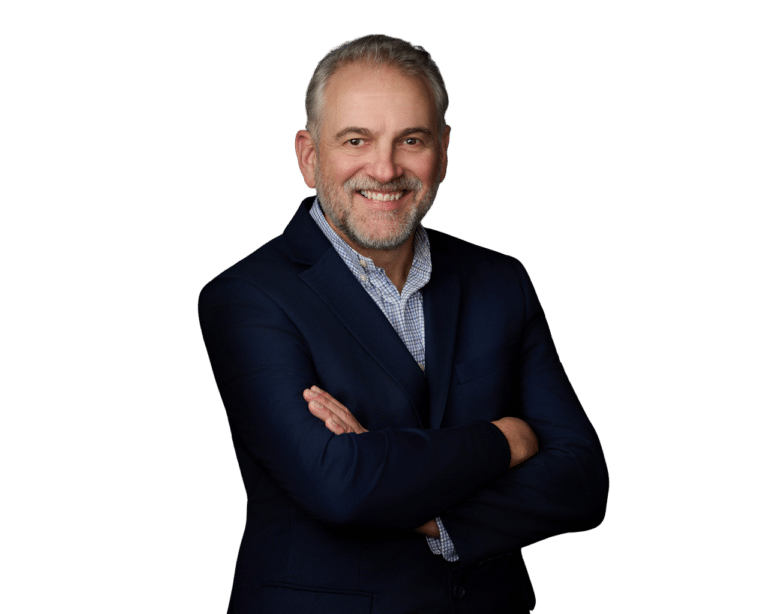 Smiling middle-aged man with short gray hair and beard, wearing a navy blazer and checked shirt, standing with arms crossed against a plain white background.
