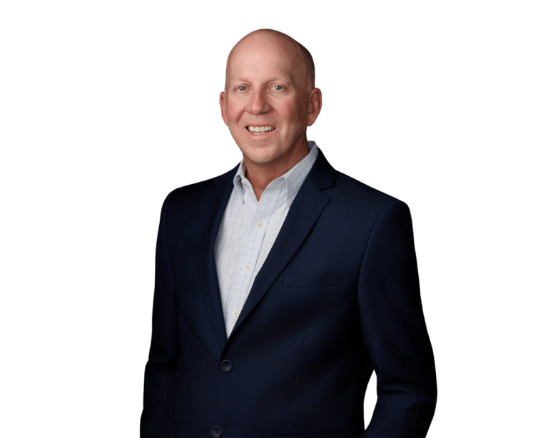 Bald man in a dark suit jacket and light dress shirt stands facing forward, smiling, against a plain white background.