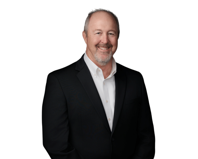 A middle-aged man with short gray hair and a beard, wearing a black suit jacket and white shirt, stands smiling against a plain white background.