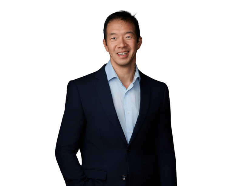 Man in a navy suit and light blue shirt, standing against a plain white background, smiling with one hand in his pocket.