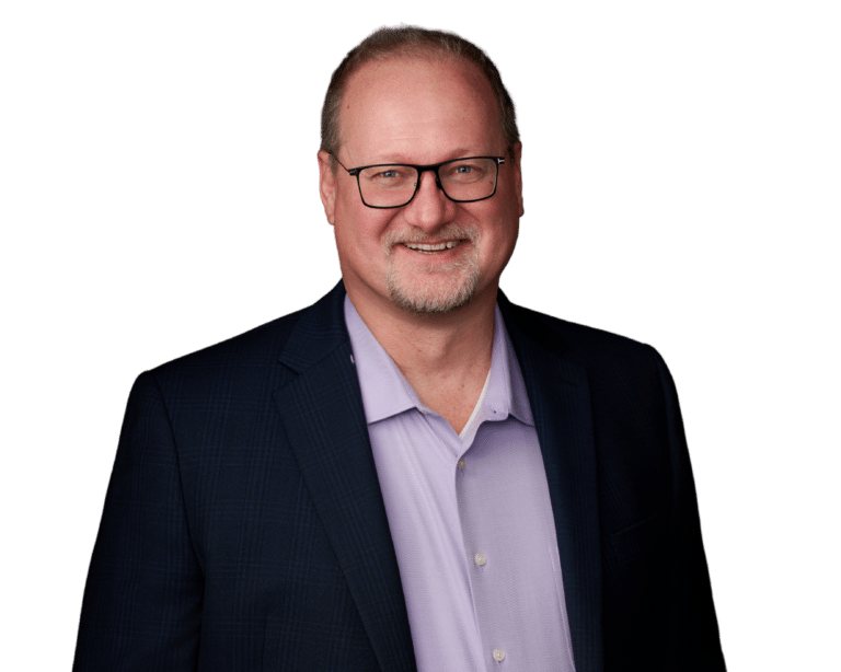 A middle-aged man with glasses, short light hair, and a trimmed beard is wearing a dark blazer over a light purple shirt, smiling at the camera against a plain white background.