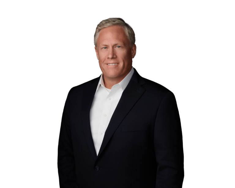 Man in a dark suit and white shirt stands against a plain white background, smiling at the camera with hands at his sides.