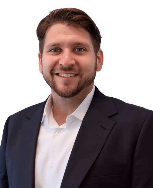 Man with short brown hair and beard, wearing a dark suit jacket and white shirt, smiling at the camera against a plain white background.