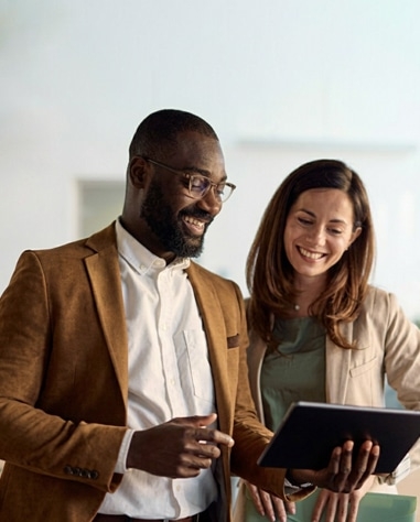 Two professionally dressed people stand together smiling while looking at a tablet device in an office setting.