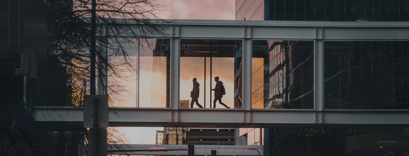 Two people walk in opposite directions inside a glass pedestrian skywalk between modern office buildings at dusk.