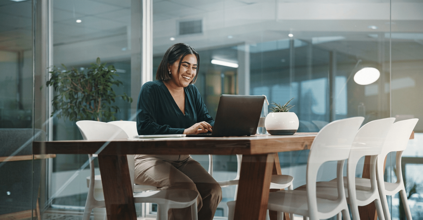 A person sits at a conference table, smiling while working on a laptop in a modern office with glass walls and a potted plant.