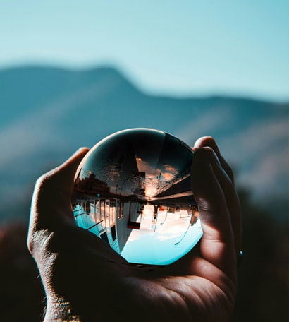A hand holds a glass sphere that reflects an upside-down image of mountains, sky, and buildings in the background.