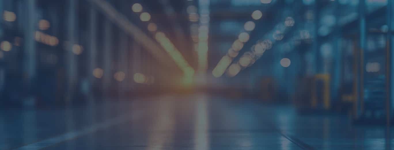 A blurred view of a large industrial warehouse with high ceilings, bright overhead lights, and shiny floors.