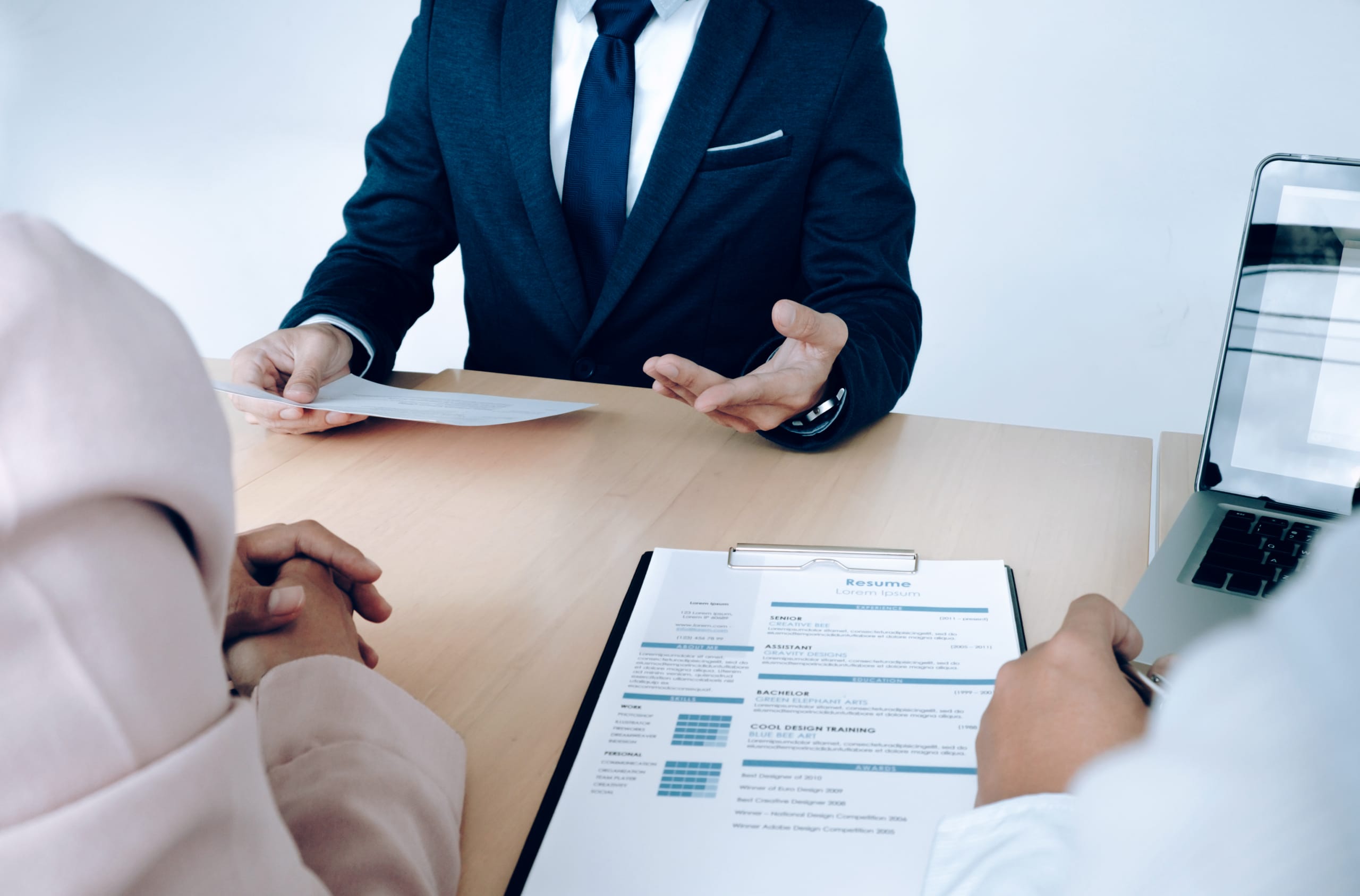 Three people sit at a table during a job interview, with one person in a suit speaking and two others holding resumes and a laptop.
