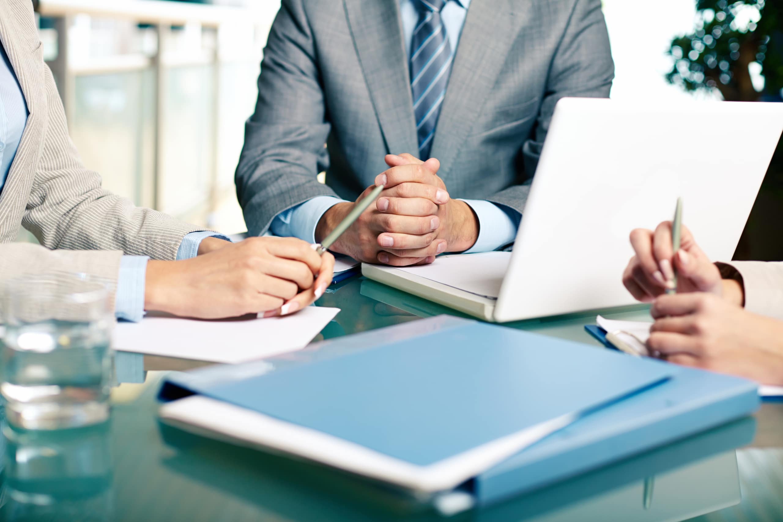 Three people in business attire sit at a glass table with documents, a laptop, and folders, appearing to have a meeting or discussion in an office setting.