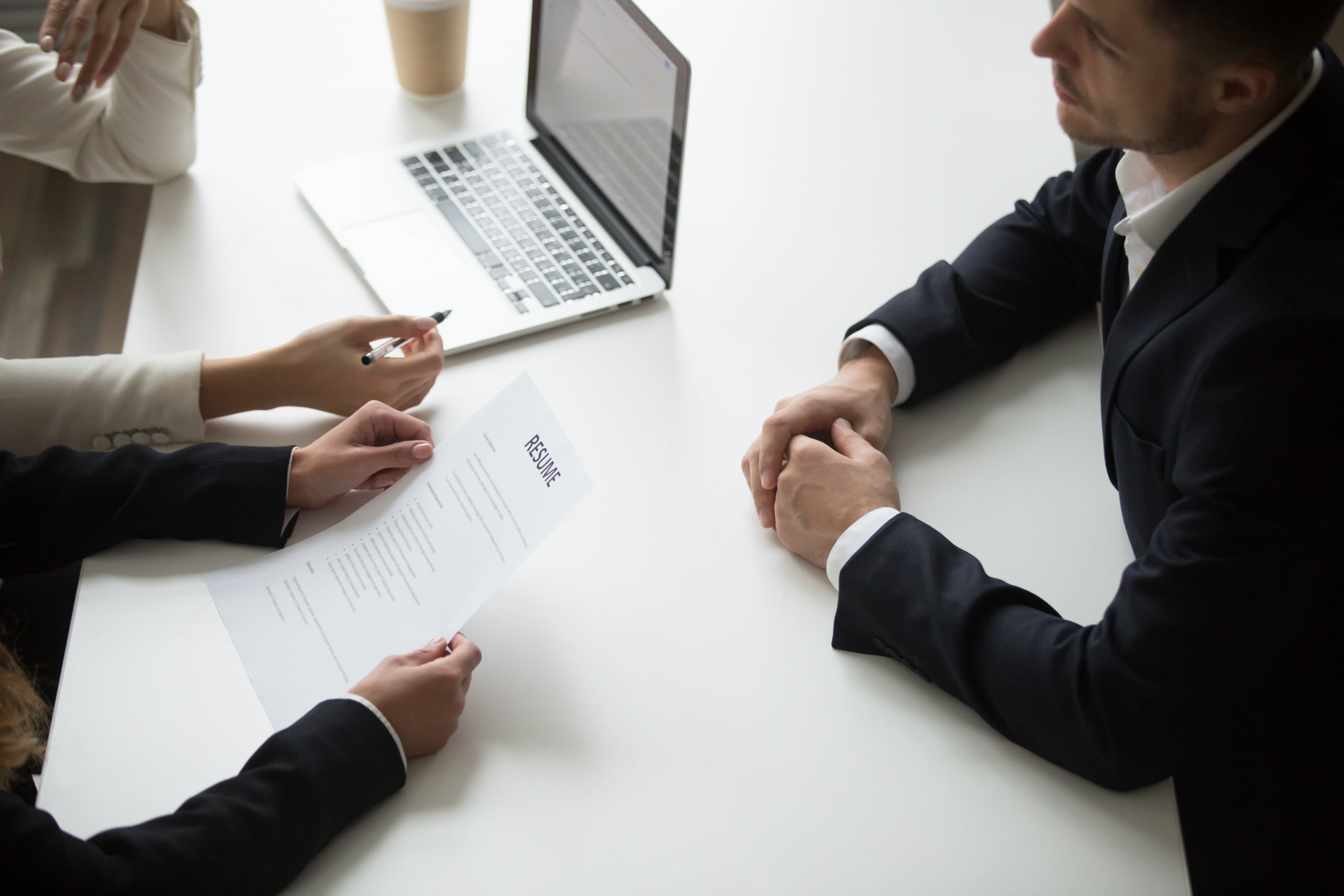 Two people reviewing a resume with a job candidate at a desk, with a laptop and coffee cup in the background.