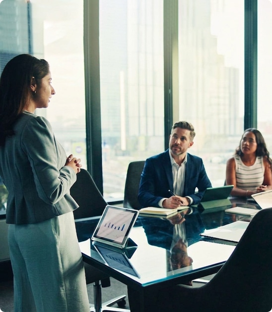 Woman presenting to two colleagues in a modern office conference room, with laptops, papers, and a cityscape visible through large windows.