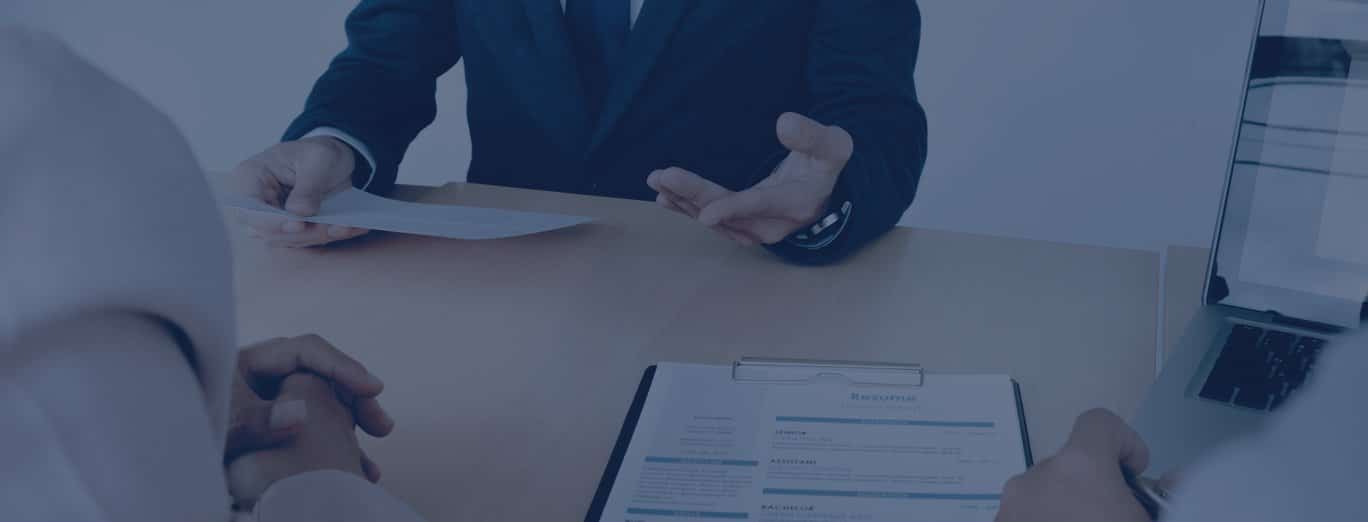 Three people sitting at a table during a job interview; one person holds a resume, another gestures while speaking, and a laptop is visible.