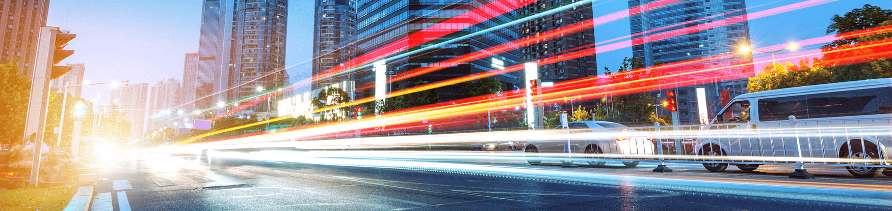 City street at dusk with light trails from moving vehicles, high-rise buildings, and stationary cars on the right side.