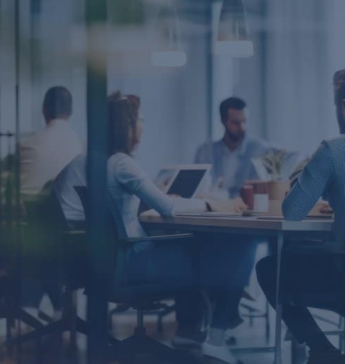 A group of people sit around a conference table in a modern office, working on laptops and having a meeting.