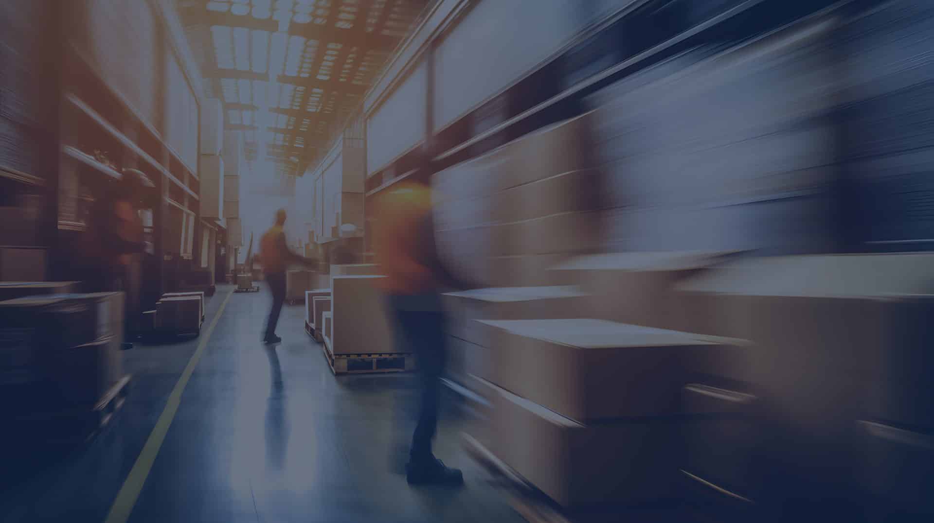 Warehouse workers moving boxes along aisles filled with stacked shelves, with motion blur indicating activity and movement.