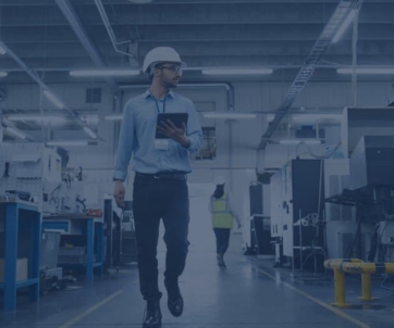 A man in a white hard hat and safety glasses walks through a factory, holding a tablet as he reviews data for a sell-side assessment. Other workers and industrial equipment are visible in the background.