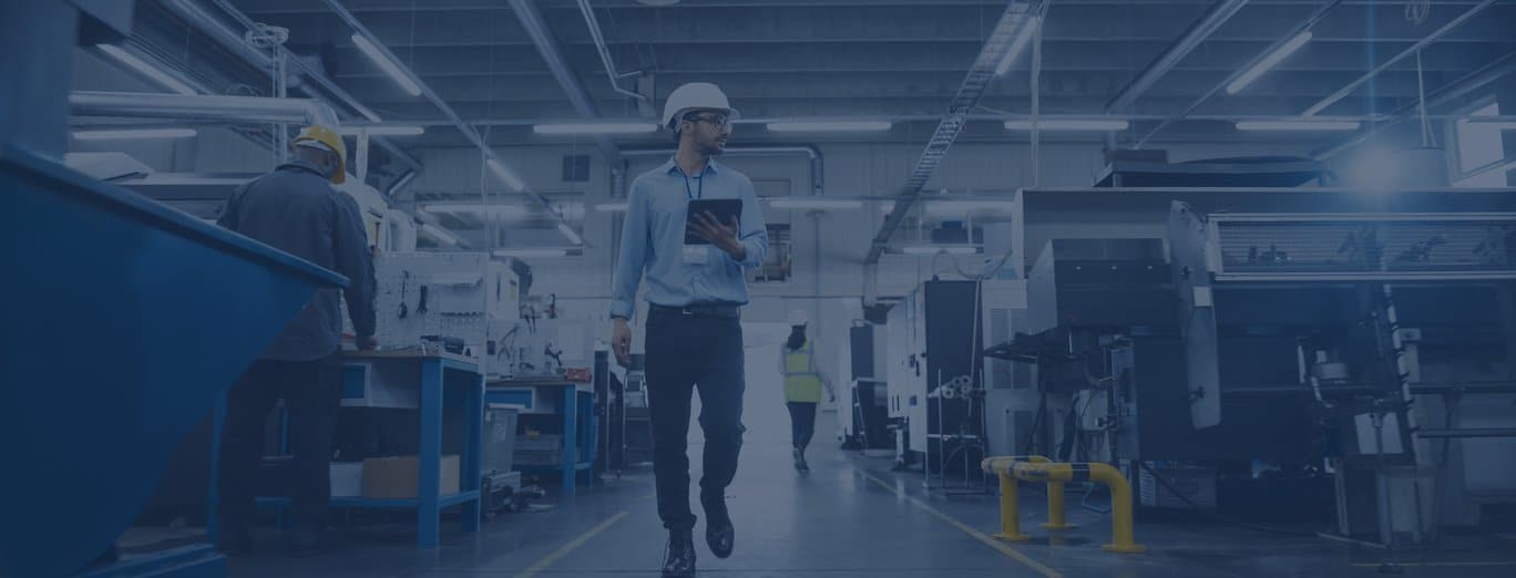 A man in a white hard hat and safety glasses walks through a factory, holding a tablet as he reviews data for a sell-side assessment. Other workers and industrial equipment are visible in the background.