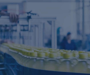 A conveyor belt moves rows of yellow-capped bottles through a factory, where workers in the background operate machinery as part of a facility that prioritizes food safety consulting standards.