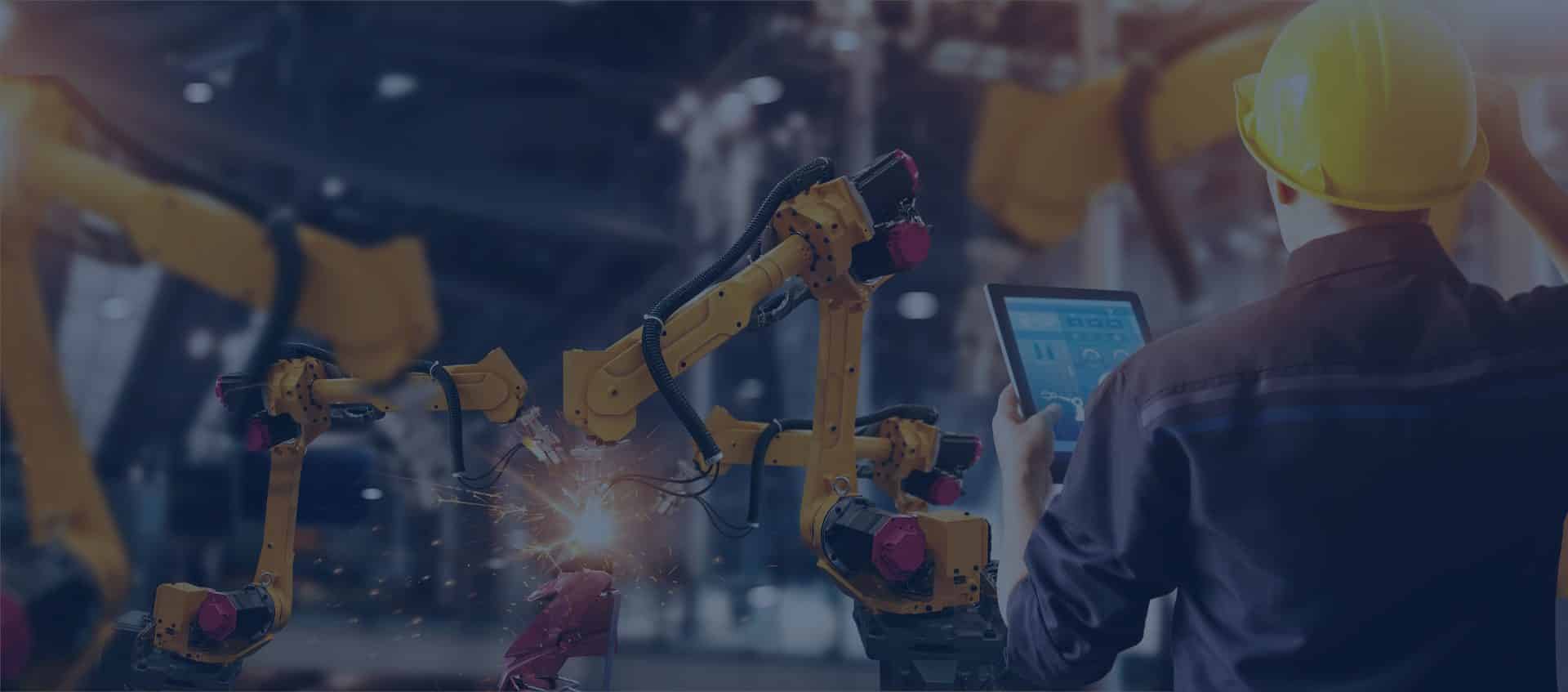 A worker in a hard hat uses a tablet to control yellow robotic arms performing welding tasks in an industrial setting.