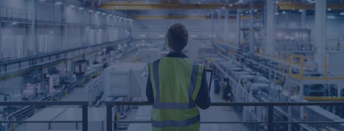 Person in a high-visibility vest stands on a platform overlooking large industrial machinery and equipment inside a factory, embodying the principles of operations improvement consulting.